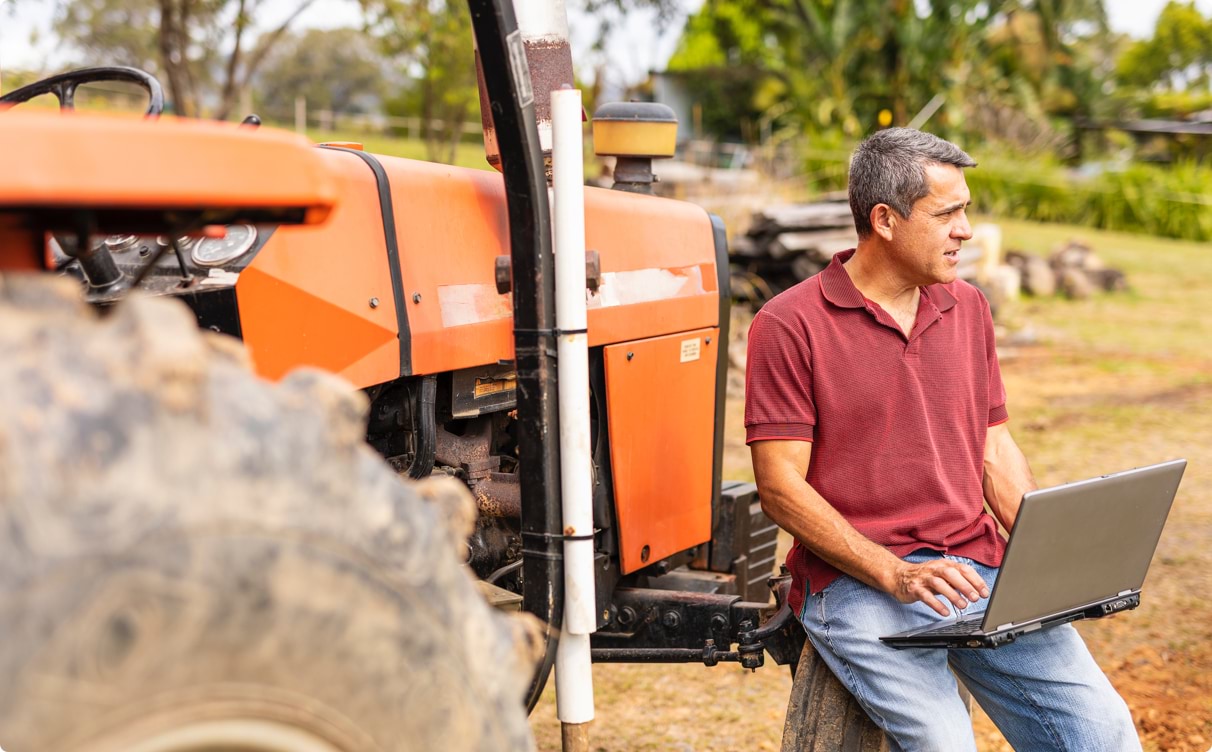 A man sitting on edge of the front wheel of a large orange tractor. He has a laptop balanced on his lap and he is looking into the distance as though he is thinking.