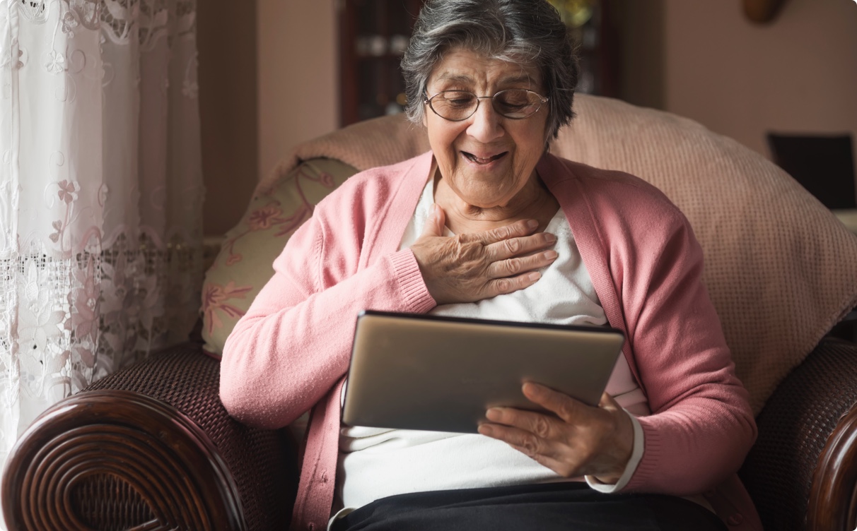 An elderly woman sits in a comfy armchair, smiling at an iPad.