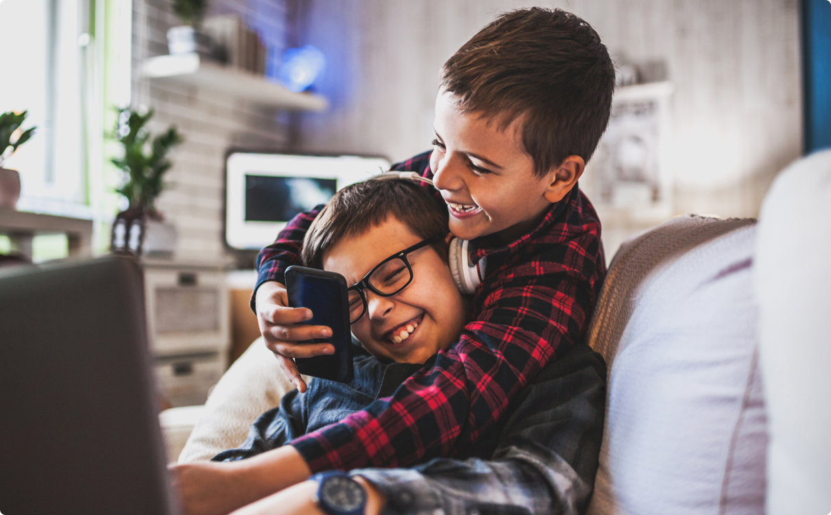 Two small boys play-wrestling on the living room sofa. One is holding a mobile phone, and there appears to be a laptop monitor out-of-focus in the foreground.