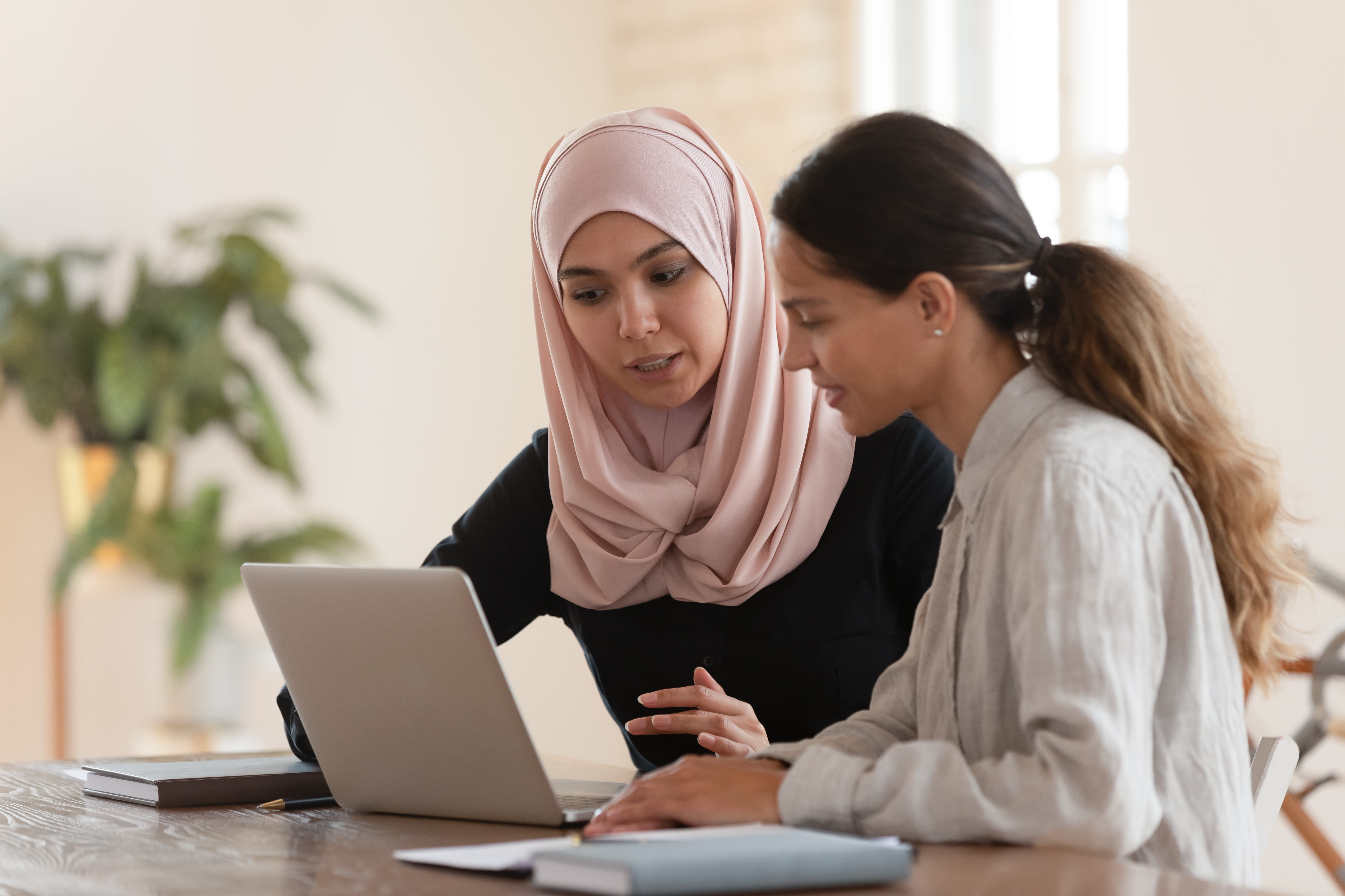 Young woman in hijab sitting with smiling colleague at table looking at computer screen