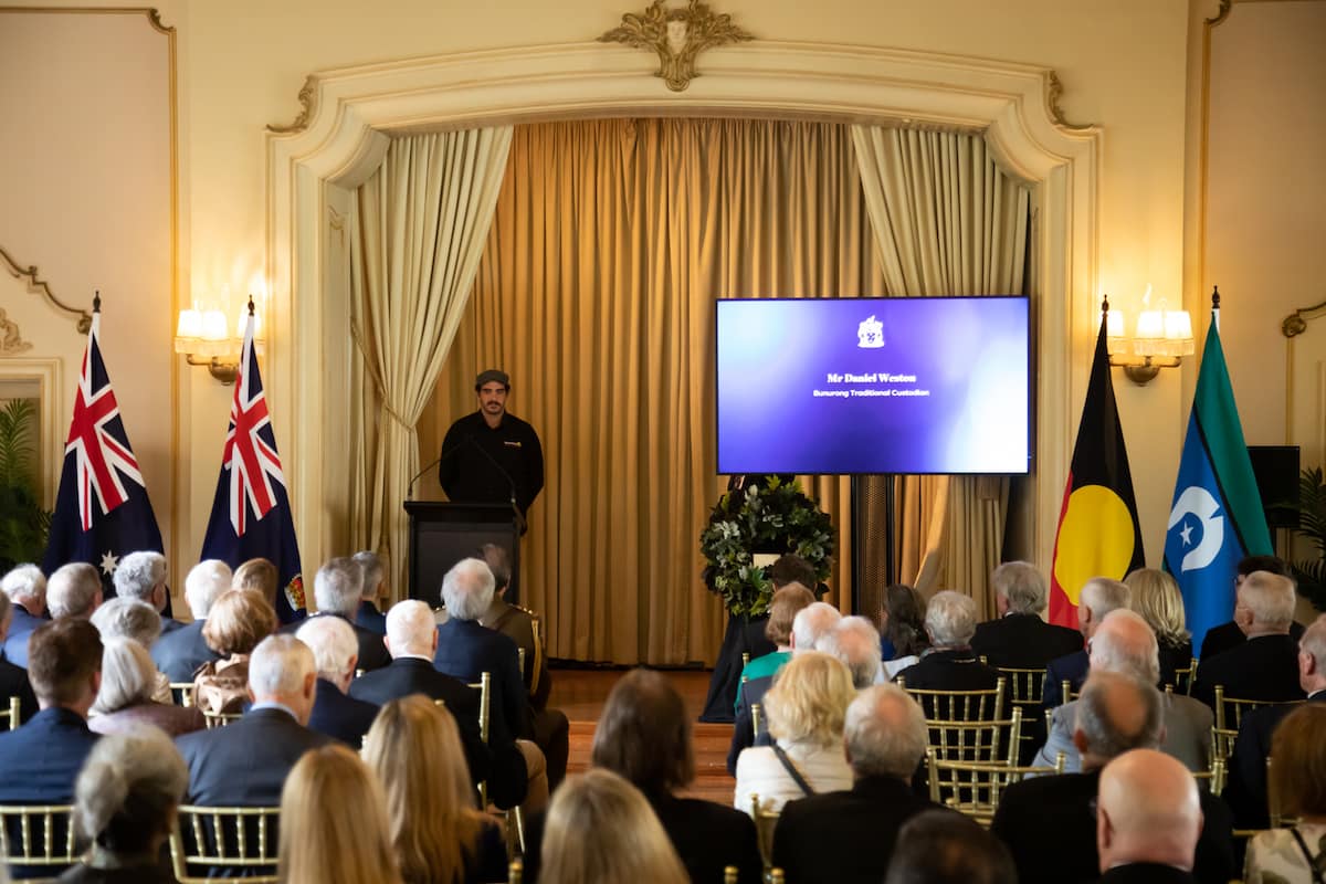 Daniel Weston stands on stage in front of a crowd, flags on either side of him