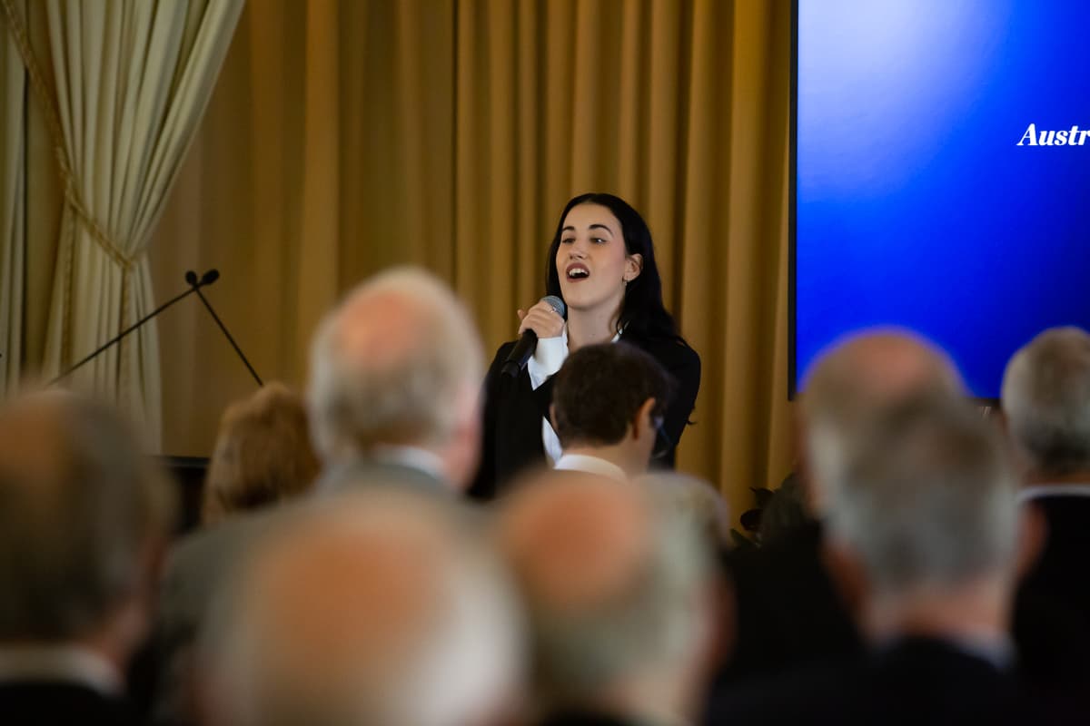 Hope Zorbas sings holding a microphone to her mouth, a crowd in front of her is standing for the national anthem