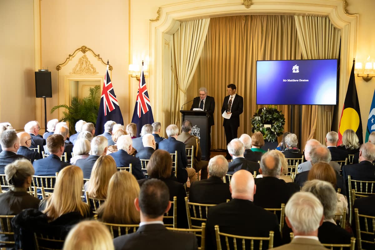 Matthew Denton stands in front of seated audience and reads at a lecturn; his brother Angus stands beside him on stage