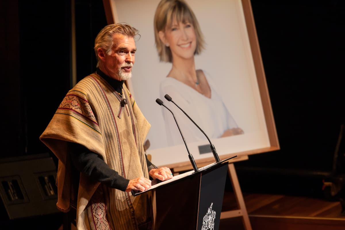 A man stands at a podium speaking into a microphone. A portrait of a woman is behind him