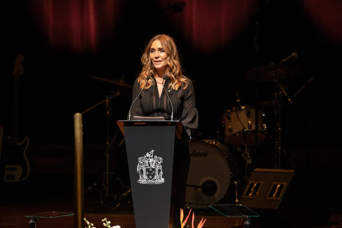 A woman with long brown hair, dressed in black stands at a podium speaking into a microphone. 