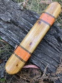 Image description: A hand crafted timber stick placed on top of grass and a log.  The timber stick has been painted with orange and black stripes at both ends of the stick