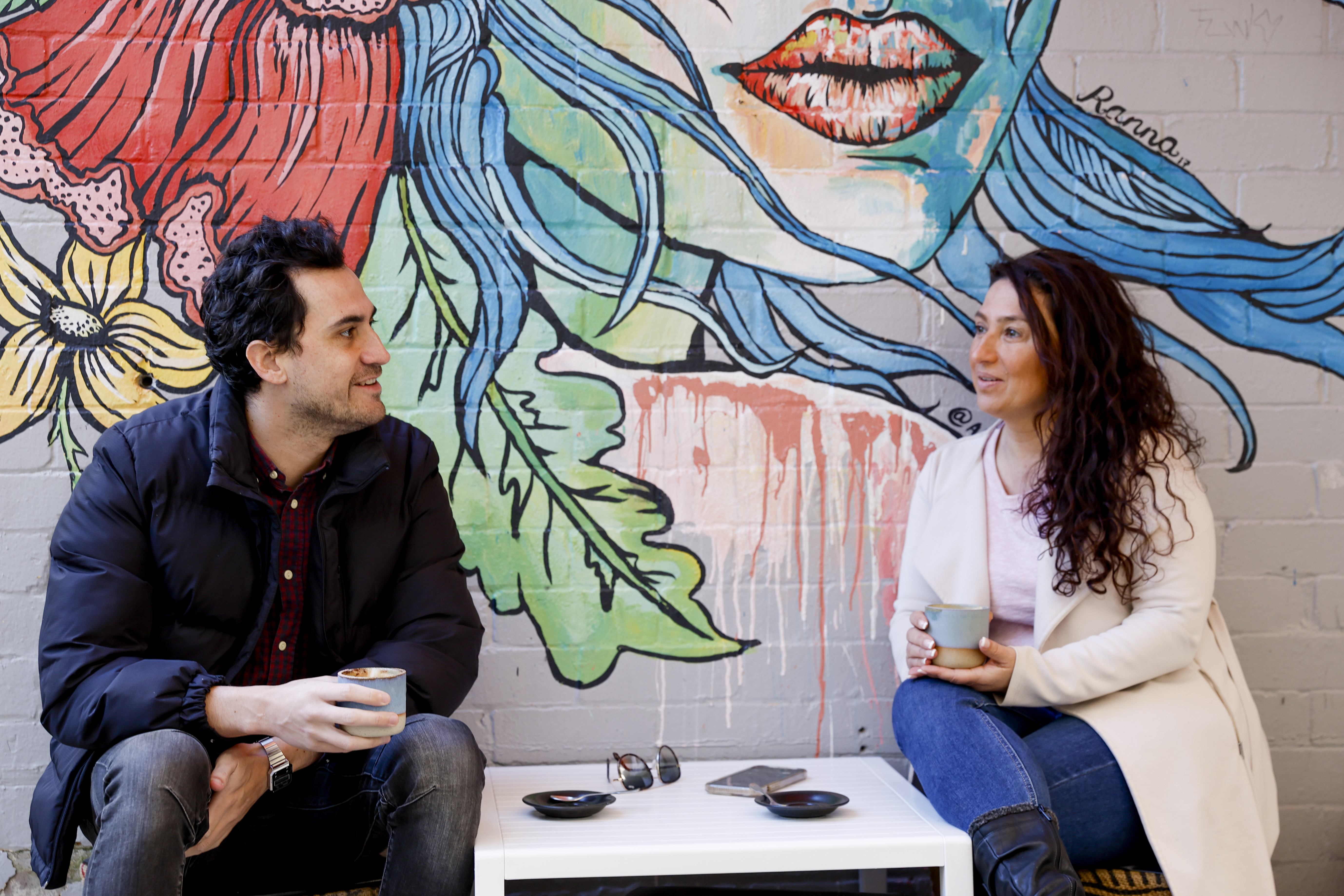 A man and a woman sit opposite each other drinking coffee with a bright mural behind them