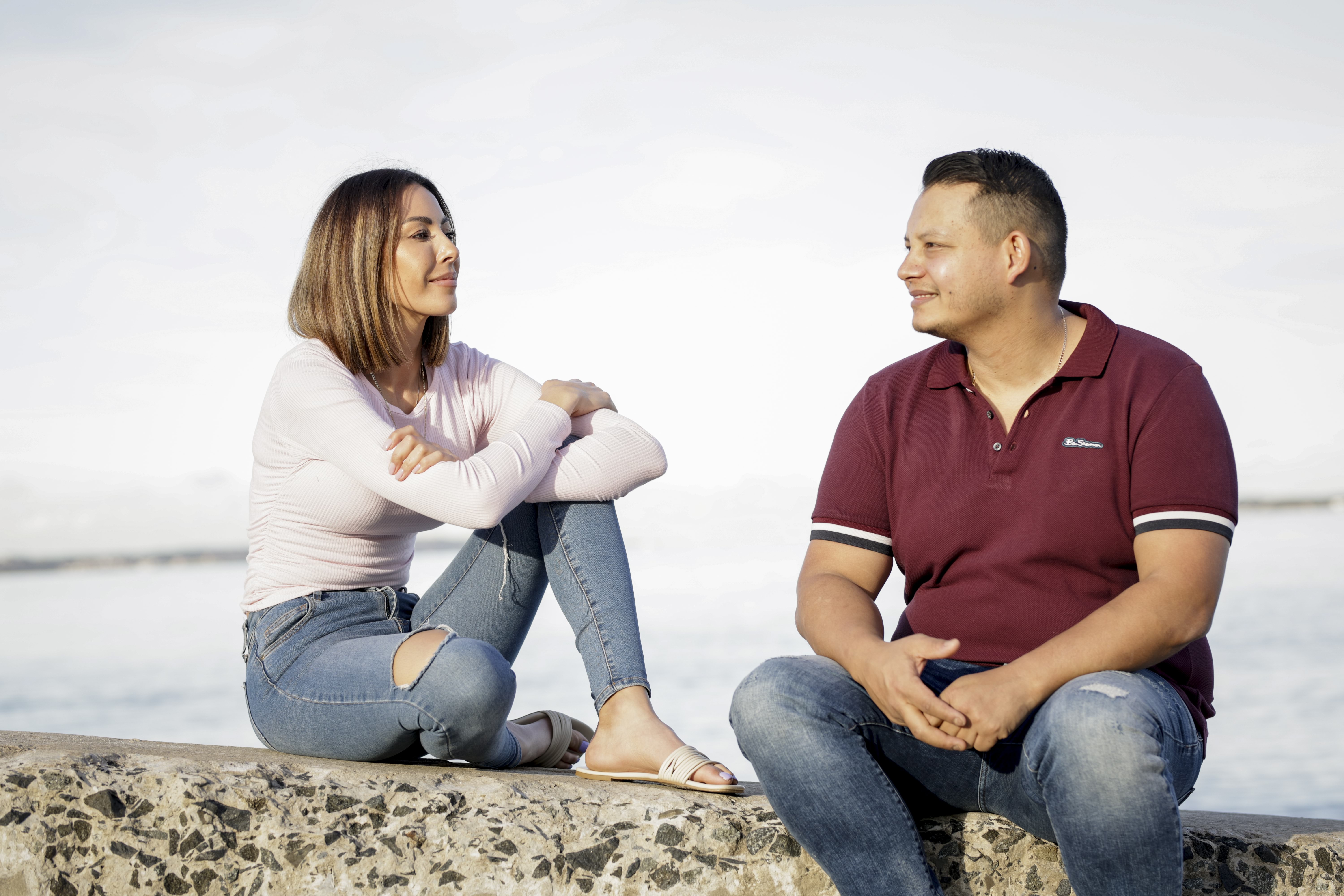 A woman and man sit on a wall next to water and smile