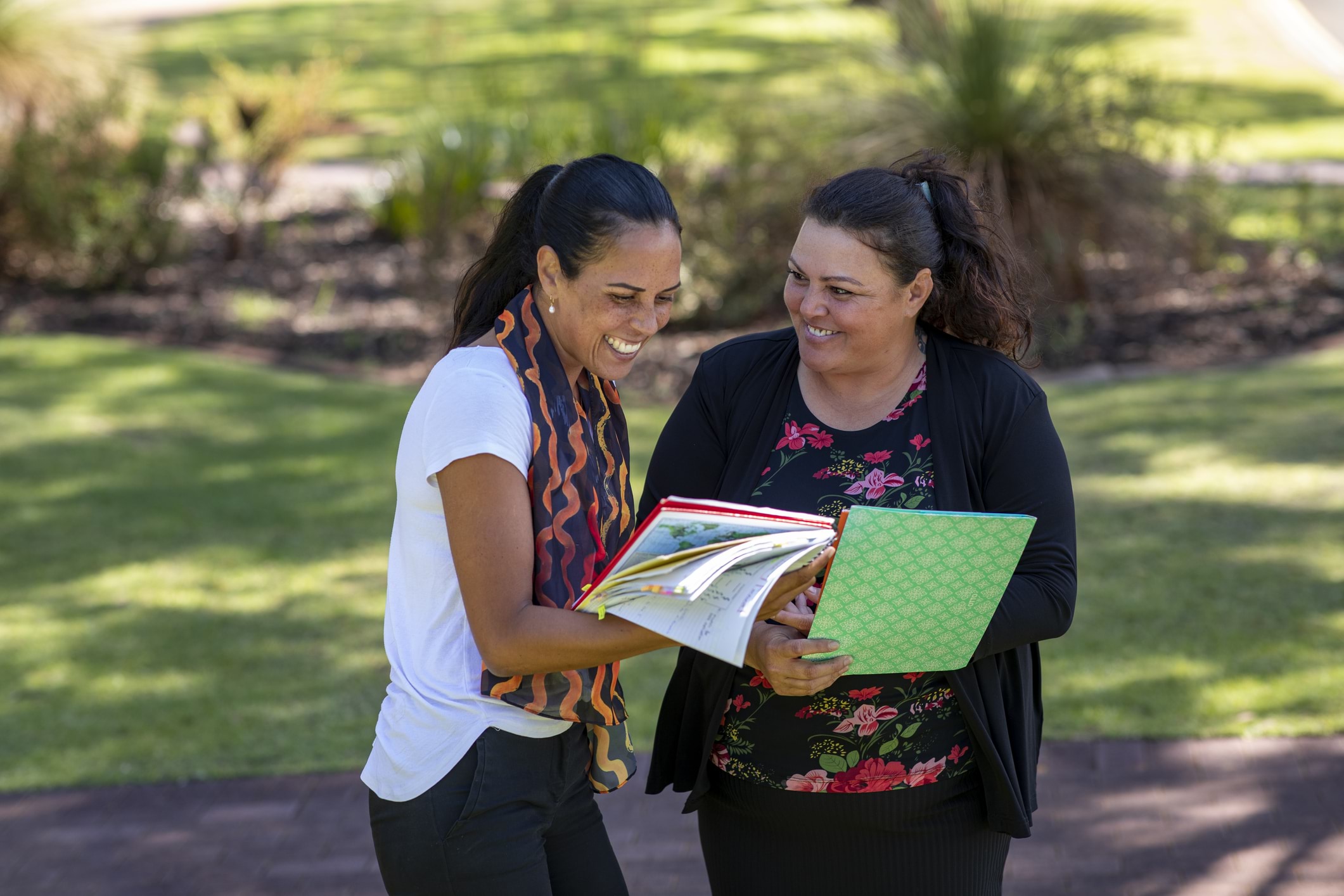 Two women stand together outside in a garden with dappled sunlight, comparing notebooks and having a friendly conversation.