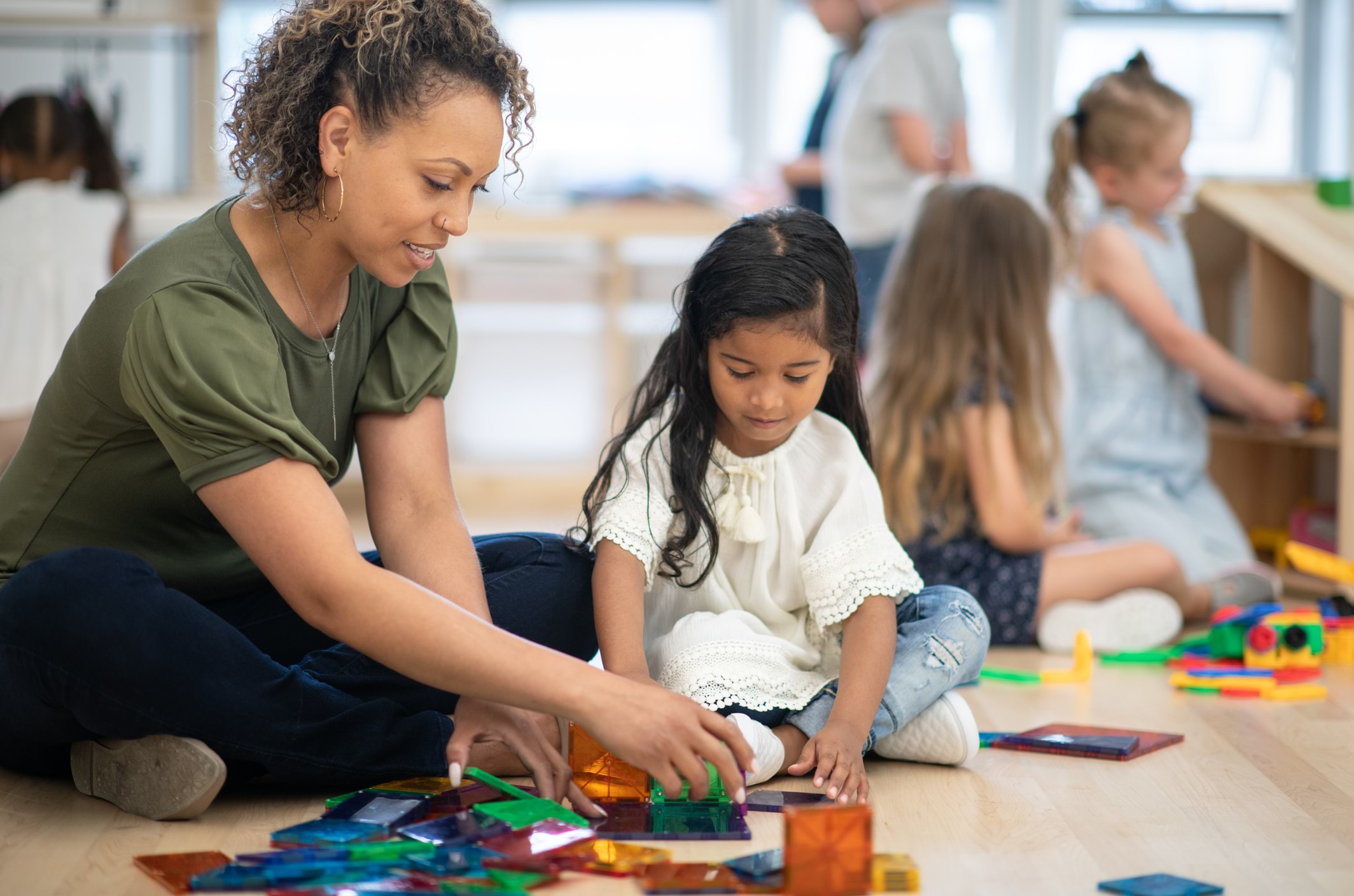 An early childhood professionals sits on the floor with a young child, both focusing intently on arranging colourful puzzle pieces. Other children play quietly in the background.