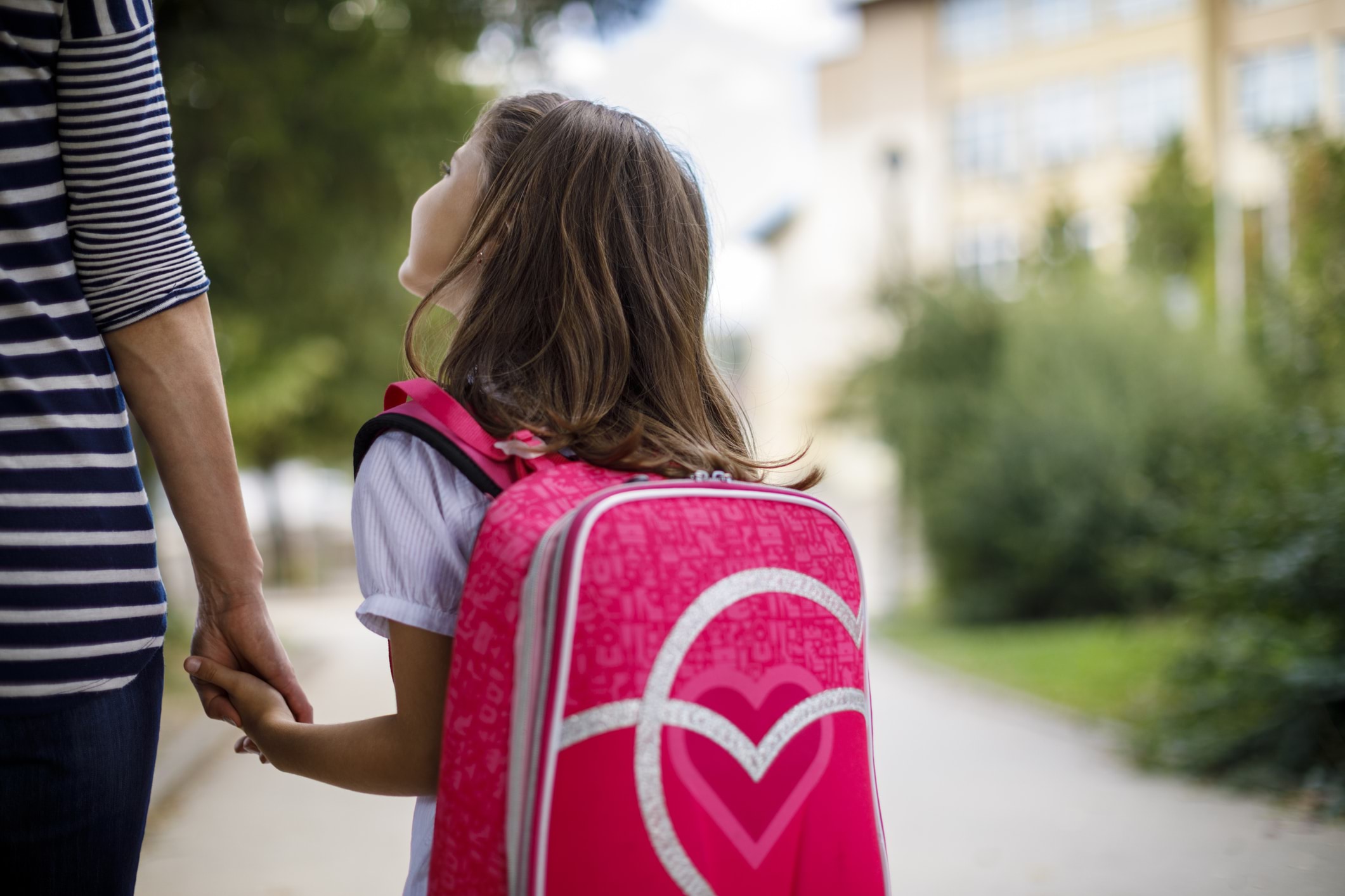 A young child with a big pink backpack holds her parent's hand as the walk toward the school gates.