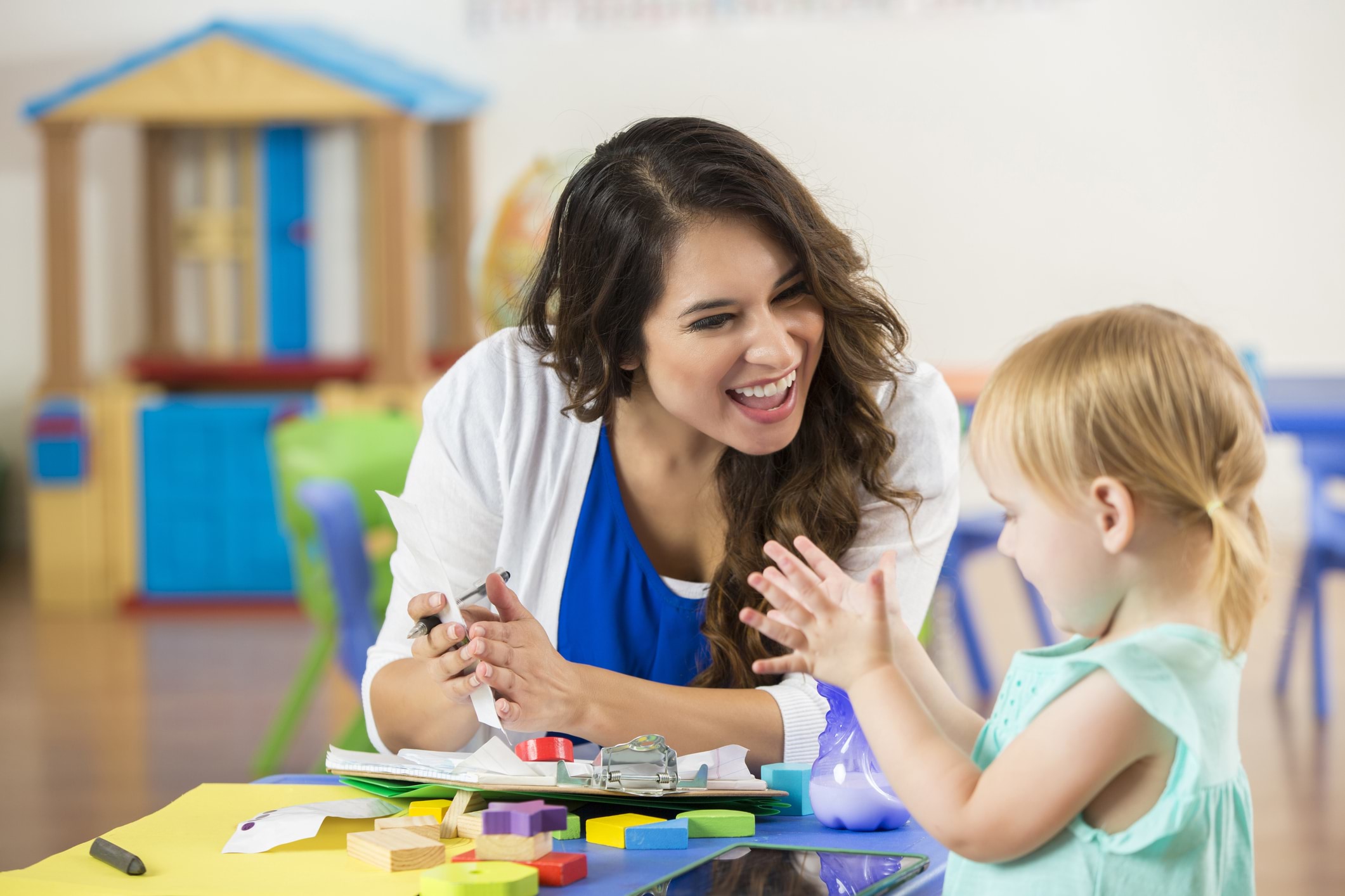 An early childhood professional sits at a table with a young child in an early childhood centre, both smiling while the child claps.