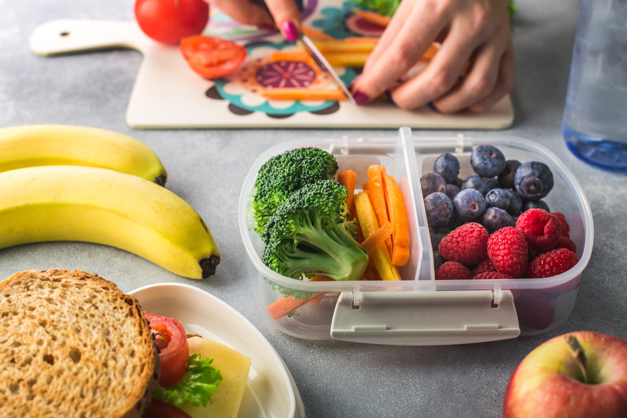 A close-up of a benchtop with a container of chopped up vegies and berries, surrounded by apples, bananas, wholegrain sandwiches, and someone's hands chopping up carrot sticks in the background.