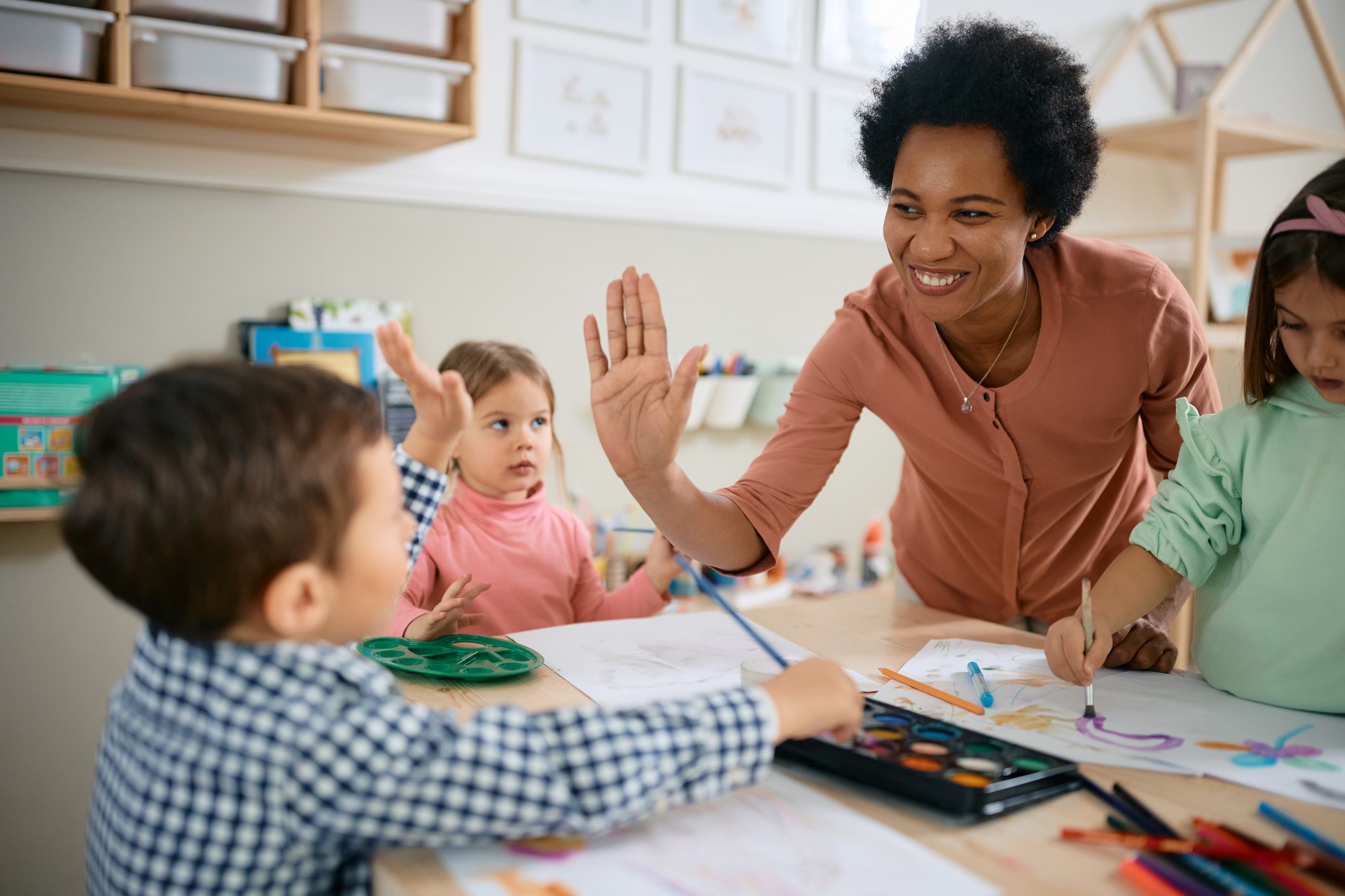 An early childhood educator high-fives a young child from across the table, while children around them paint with watercolours.