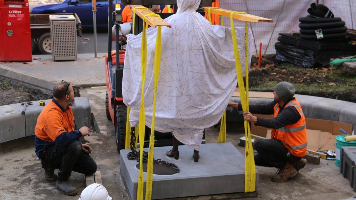 Two workers in orange high vis vests communicate to the forklift driver that the statue is in place.