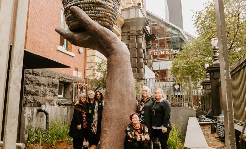 Creative Resilience artwork. A sculpture of a forearm and hand holding a basket. Located on Lonsdale Street outisde the Queen Victoria Womens Centre. Six member of the Ngardang Girri Kalat Mimini artists collective are gathered around the base of the scuplture.