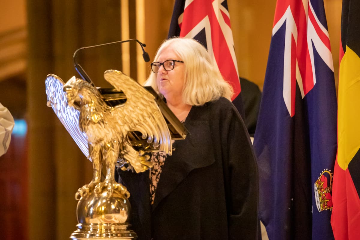 Aunty Di Kerr OAM speaks at a podium shaped like an eagle. There are flags behind her.