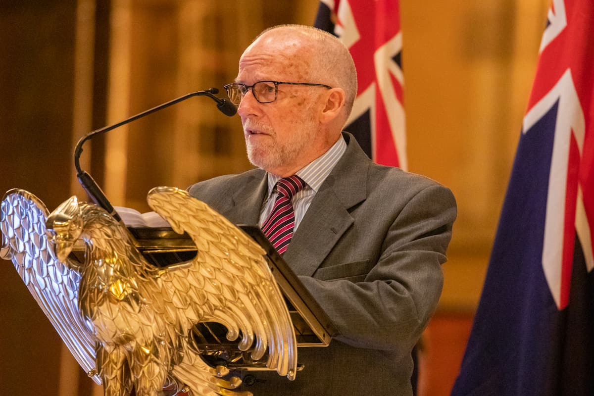 Frank O'Connor is dressed in grey and wears glasses. He speaks at a gold podium shaped like an Eagle. 