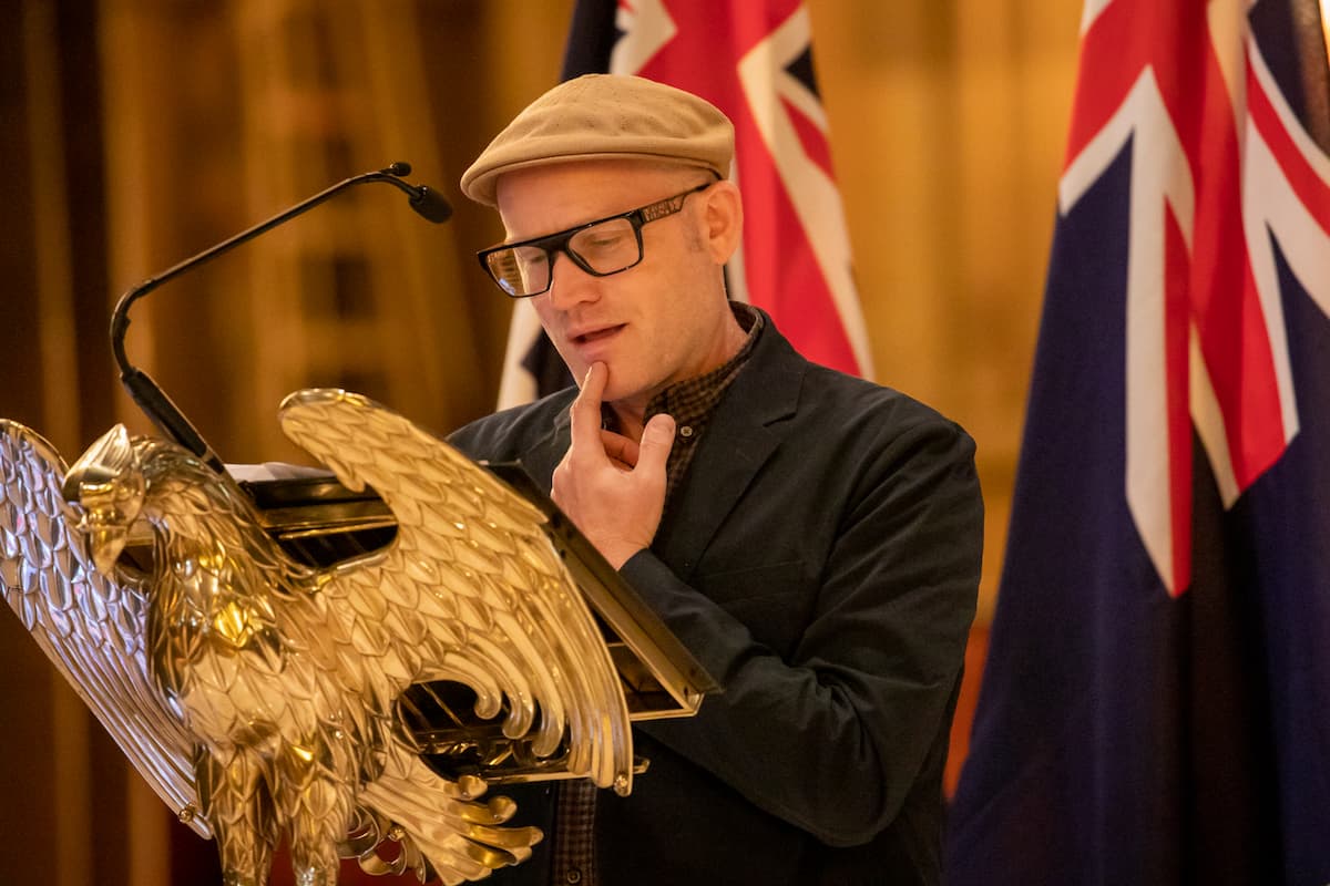 John Safran is speaks at a gold podium shaped like an eagle. He wears a beige flat cap and black glasses. He is touching his chin while he speaks.