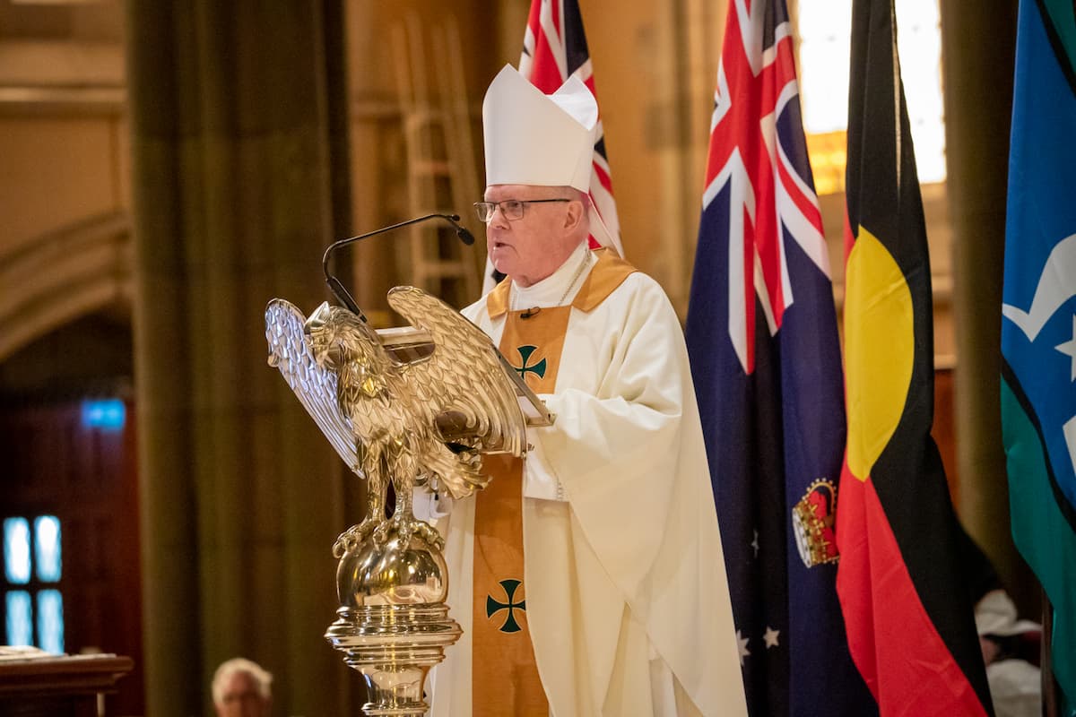 The Archbishop of Brisbane is dressed in white and gold robes and wears a white mitre on his head. He speaks at a podium in the shape of a golden eagle.