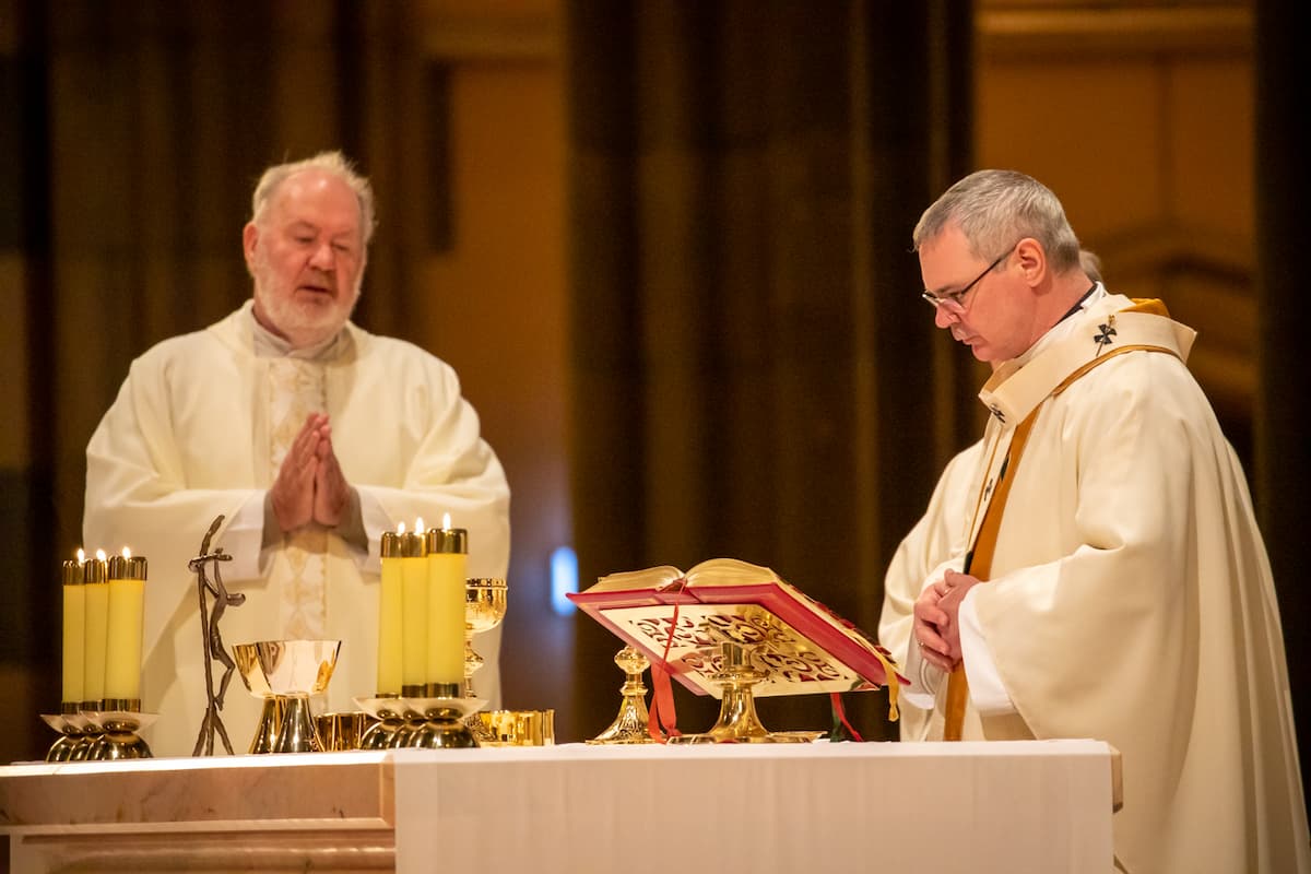 Two priests stand at the altar in prayer, dressed in white robes.