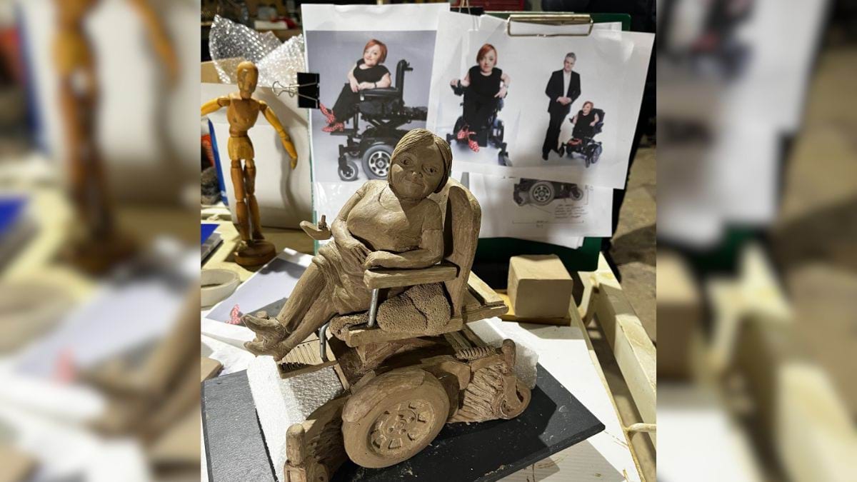 A small clay sculpture of Stella Young. On the wall behind are photos of Stella, which are the inspiration for the sculpture.