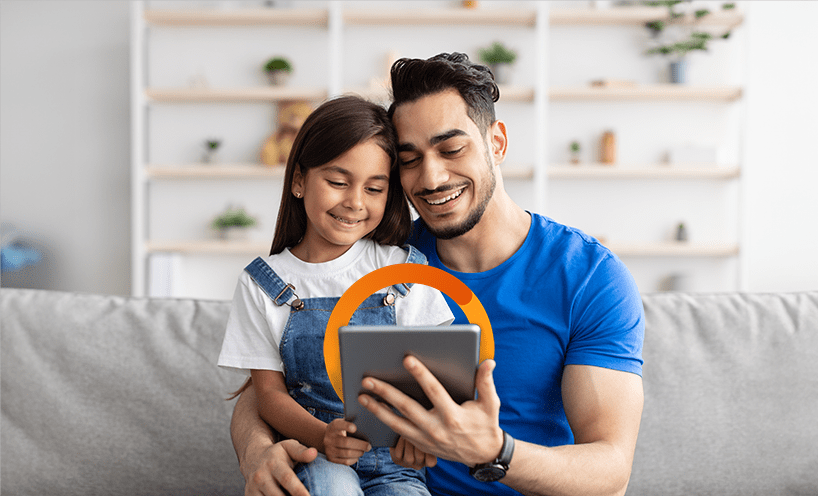 Father has young daughter on his lap, they are smiling and looking into an ipad