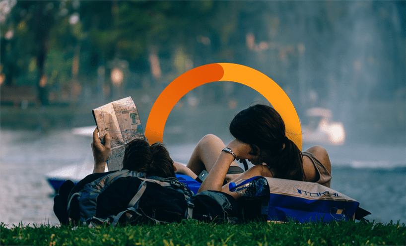 A couple are lying on the grass in front of water, looking at a map