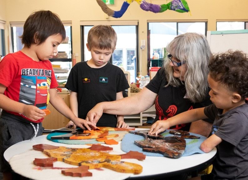 Three young children and an older woman sit at a table doing puzzles.