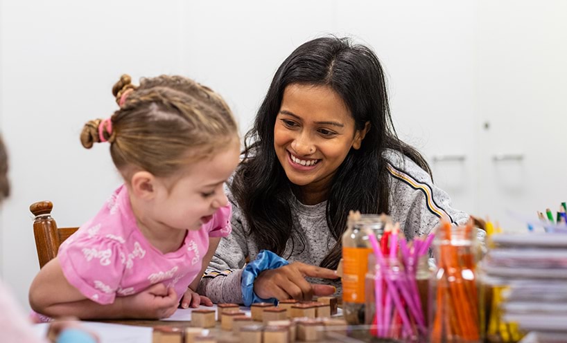 A kinder teacher sits with a kinder students. They're smiling at each other as they work on a puzzle together.