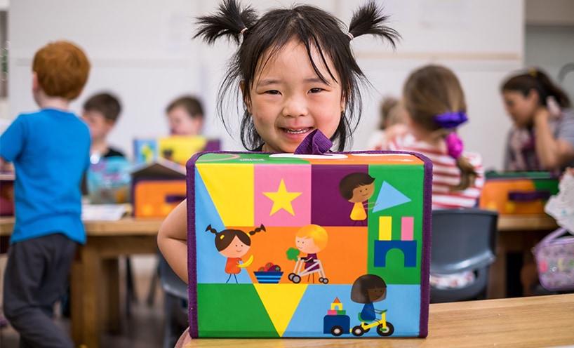 Decorative image of a kinder girl with pigtails smiling at the camera with her kind kit in front of her