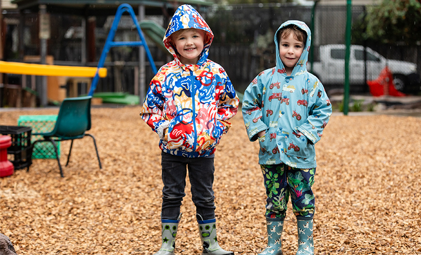 Two kindergarten children in colourful rain jackets are smiling at the camera and standing in a playground.