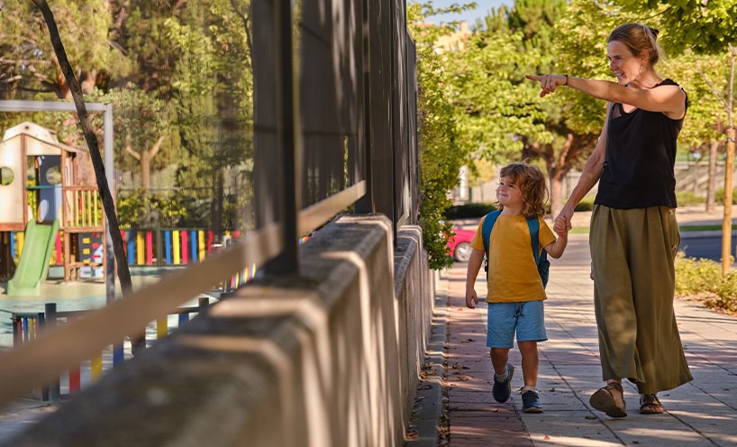 A mother and her child walk together holding hands. The mother is pointing off camera towards a playground.