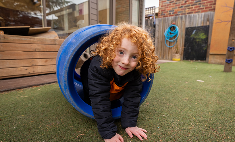 Kinder student smiles at the camera as she crawls through a tunnel.