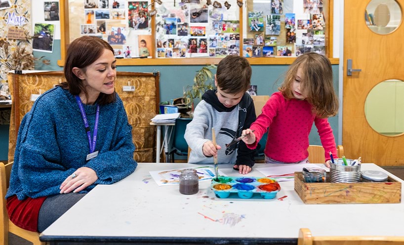 A kindergarten teacher sits with two kinder students as they paint together.