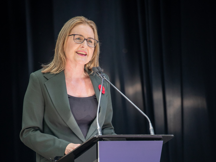 a woman in a dark green suit stands at a podium speaking to an audience