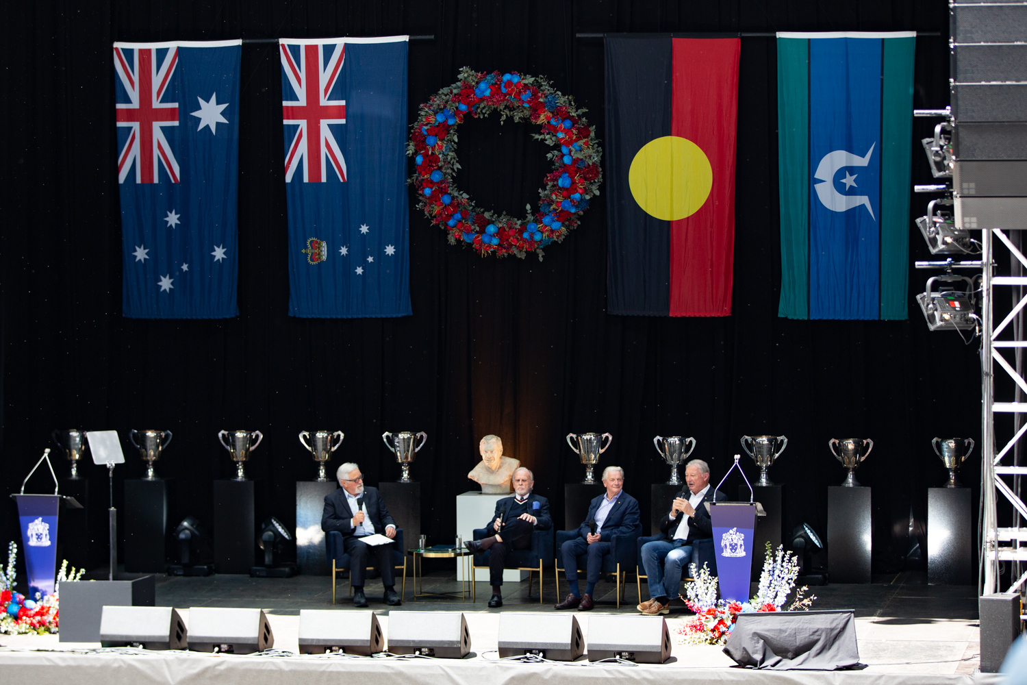 a stage is pictured that has 4 large flags draped in the background and is lined with AFL premiership cups in the background. There are four men sitting on chairs on the stage who are talking to each other and the audience through microphones 