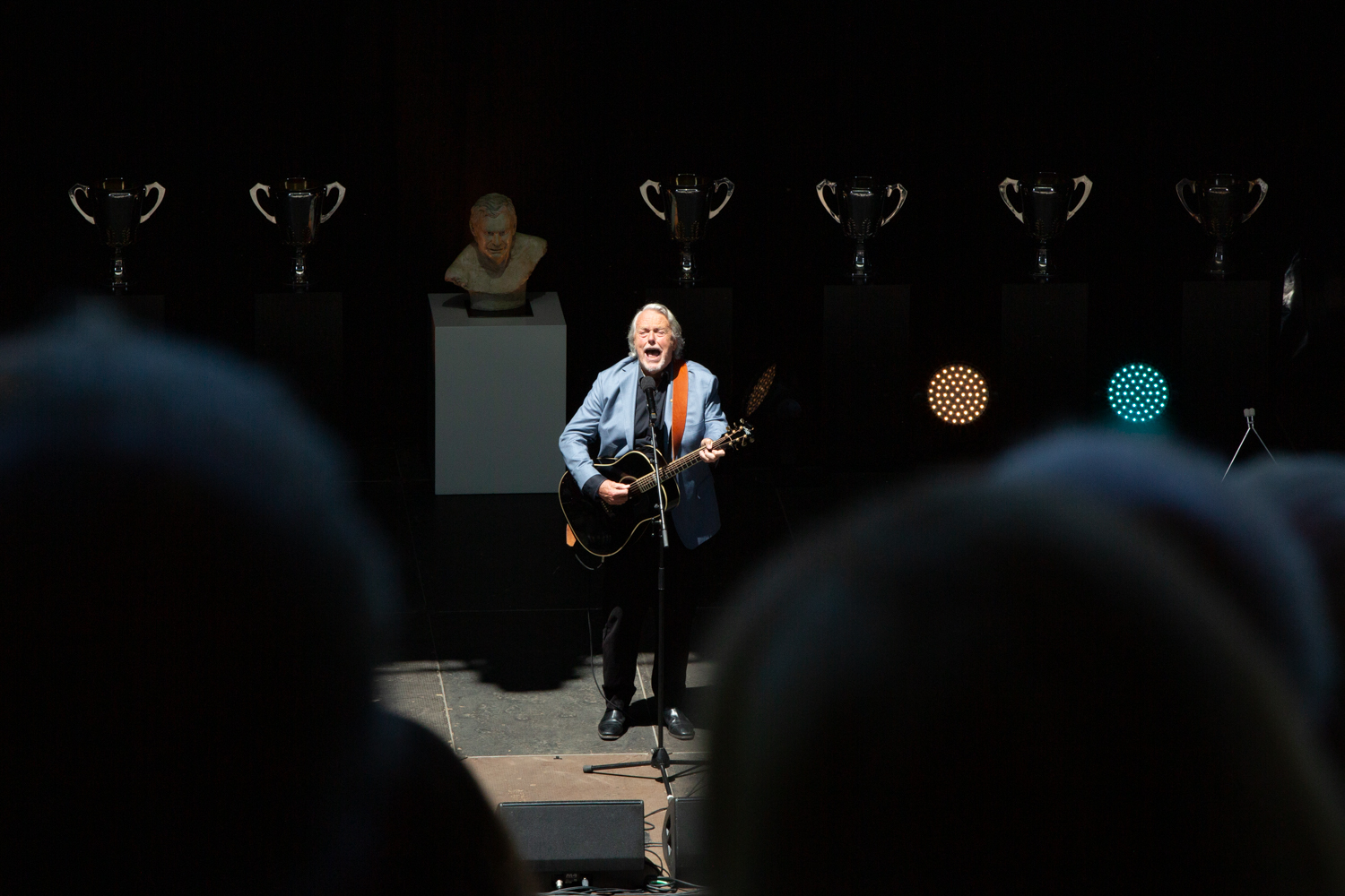 a man in a blue suit jacket is playing the guitar and is singing through a microphone on stage to an audience 