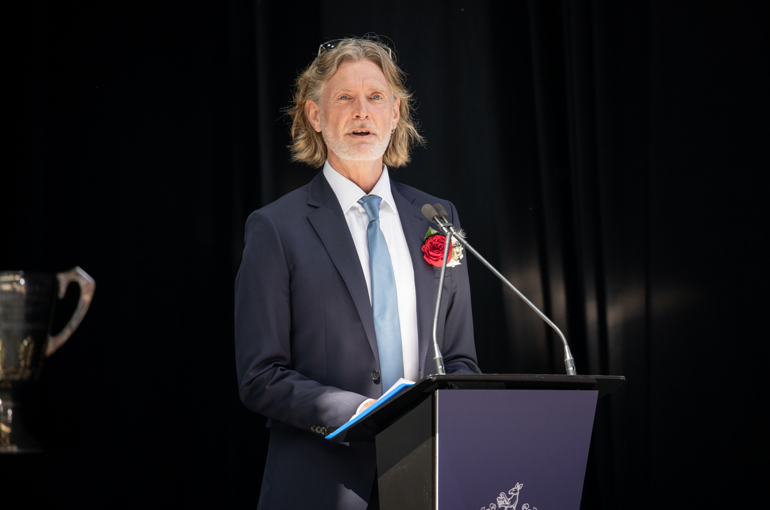 A man in a black suit with a blue tie stands at a podium speaking to an audience