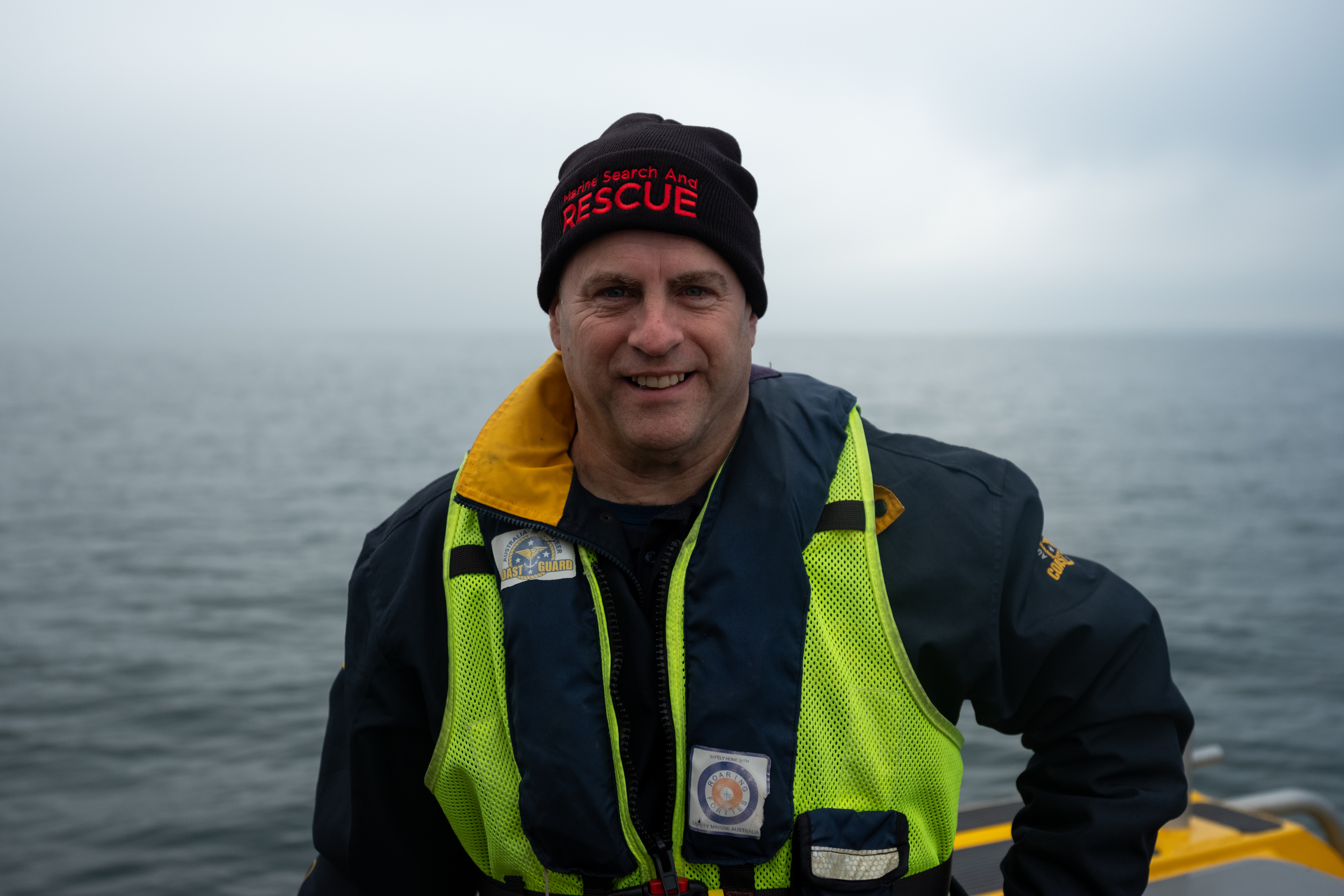 Image shows a man smiling. He is wearing a Marine Search and Rescue hat, life jacket and he is standing on a boat.