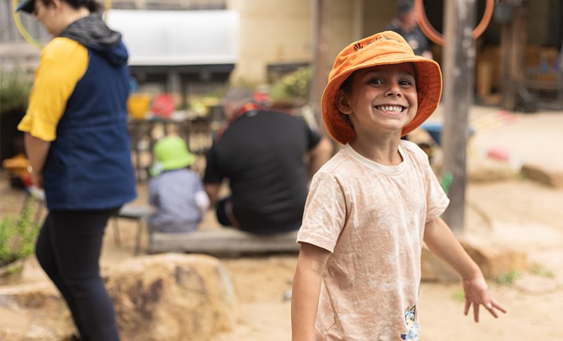 A kinder student smiles at the camera. He is outside and wearing an orange bucket hat and a cream t-shirt.