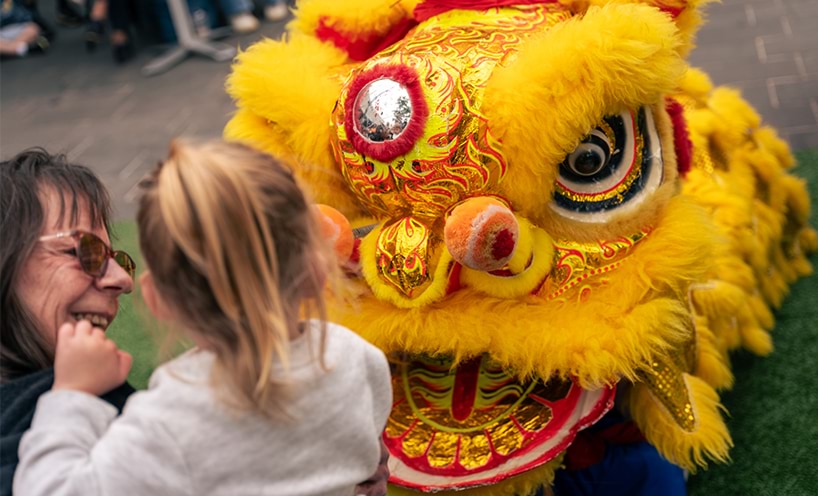 A kinder student faces the head of a decorative yellow Chinese dragon costume. She is with her kinder teacher who is smiling at her. Their backs are turned to the camera.