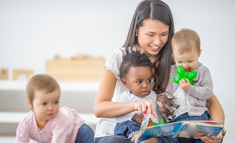 A kinder teacher is reading a picture book to three toddlers. Two toddlers sit on her lap and the other toddle is crawling behind her. They are all looking at the picture book in her hands. 