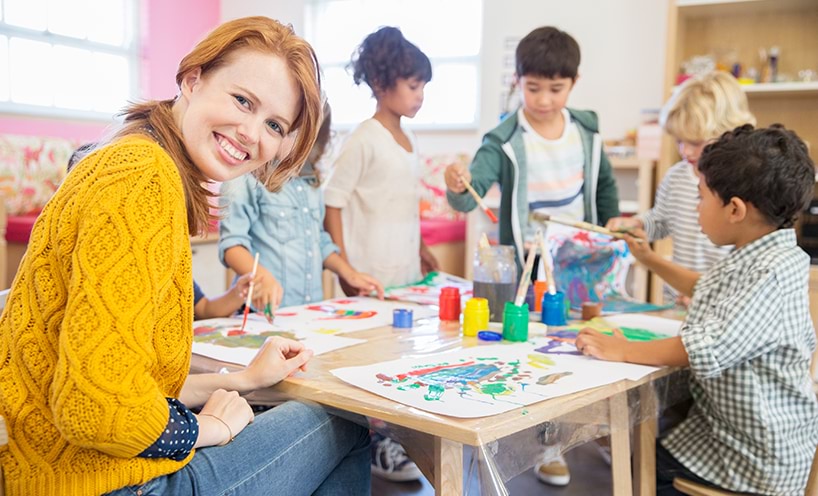 A kindergarten teacher sits with kinder students at a table. They are painting and have various paints and materials on the table. The kindergarten teacher is turned towards the camera and smiling.