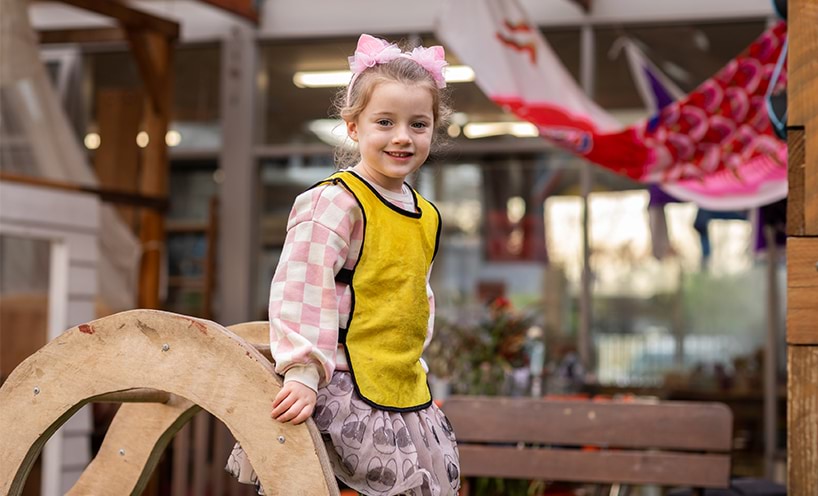 A kinder girl stands on some play equipment outside. She is smiling at the camera and wears a yellow vest over a pink jumper and skirt.