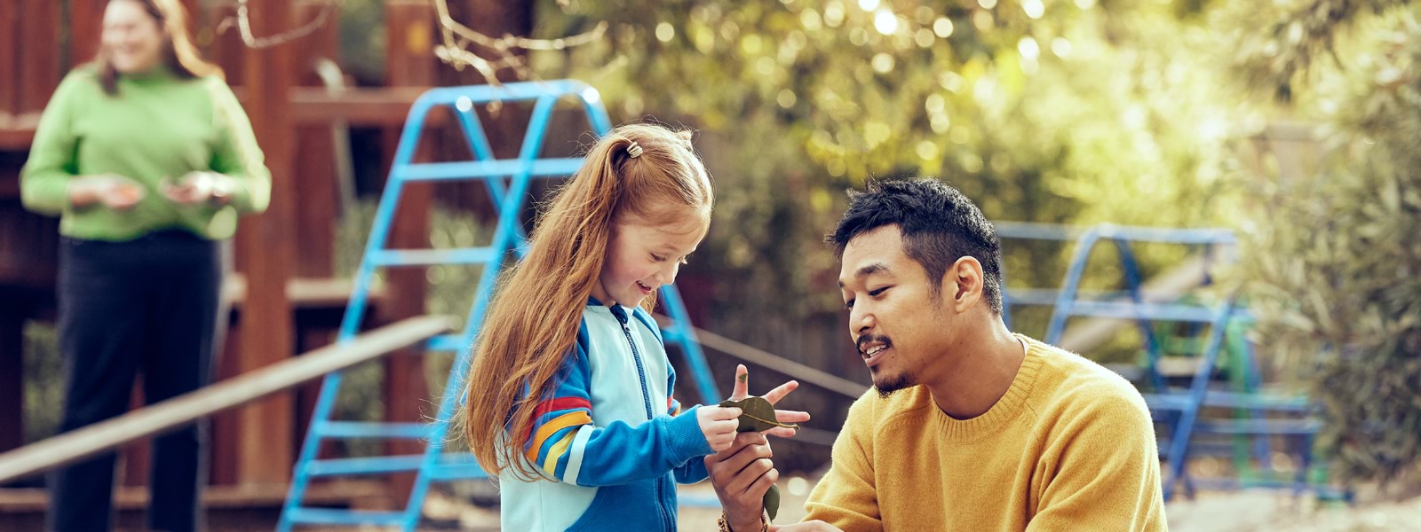 Male kindergarten teachers shows a young girl a leaf outside in a playground surrounded with trees, with early childhood educator in the background.