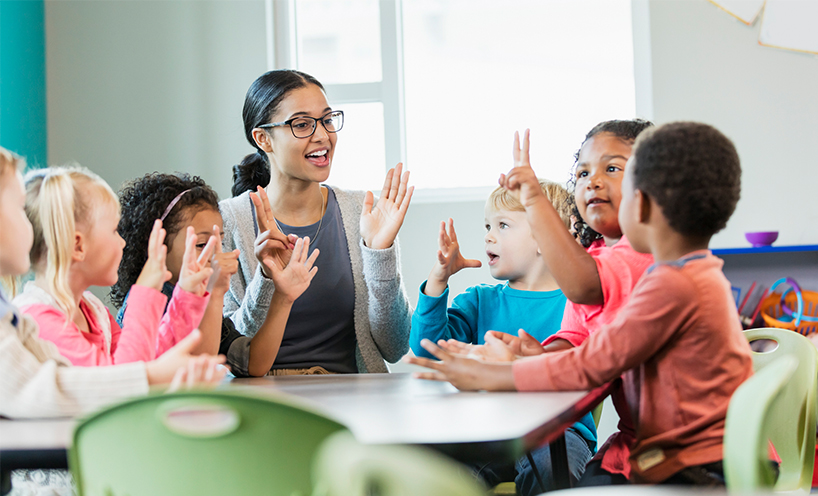 A kinder teacher sits at a table with a group of 6 children. They're all counting using their hands.