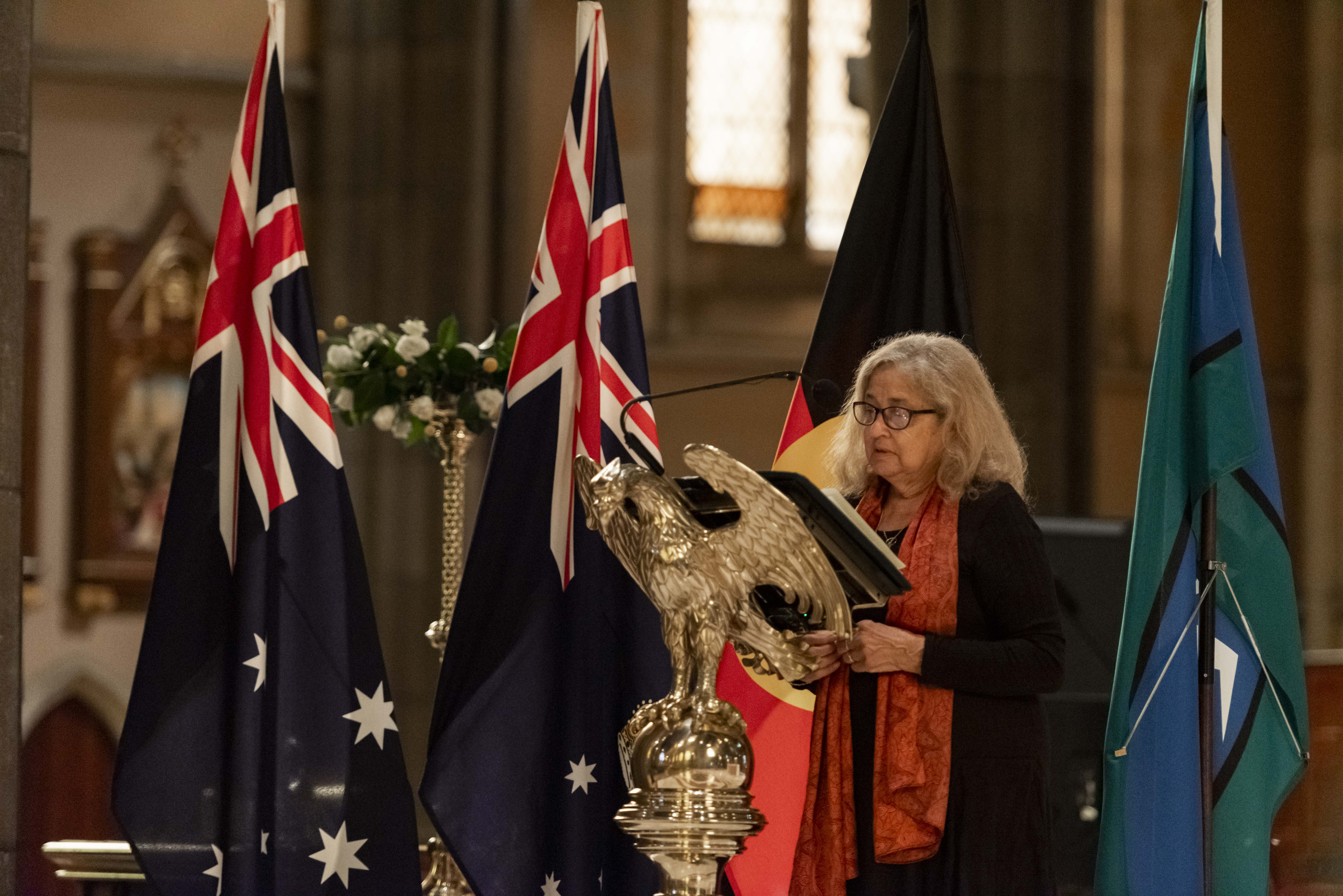 A woman with long blonde hair in a black outfit and orange scarf speaks into a microphone at a church pulpit, with the Australian, Victorian, Aboriginal and Torres Strait Islander flags in the background. 