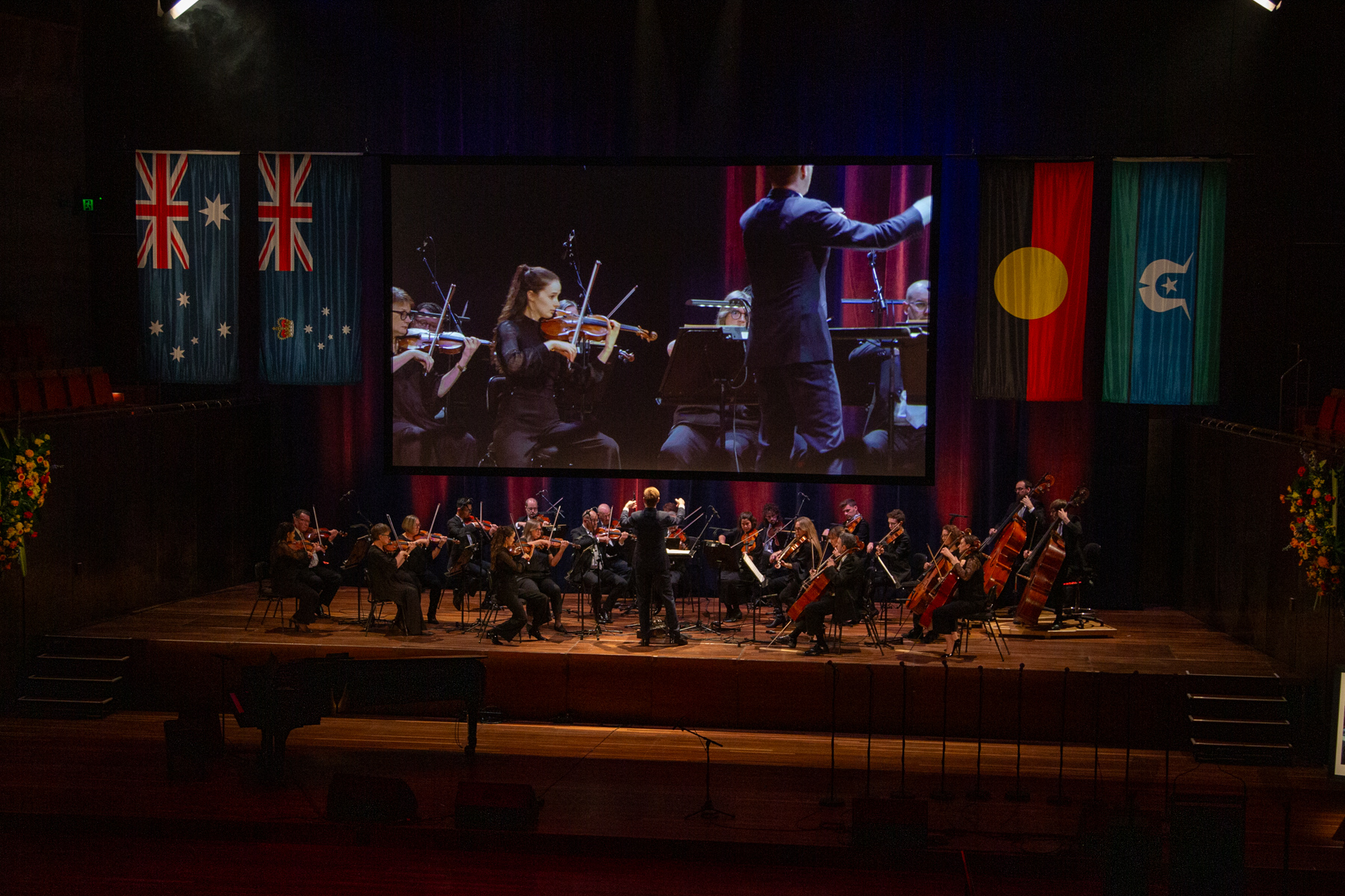 An orchestra on stage with a screen and the flags of Australia, Victoria, Australian First Peoples and Torres Strait