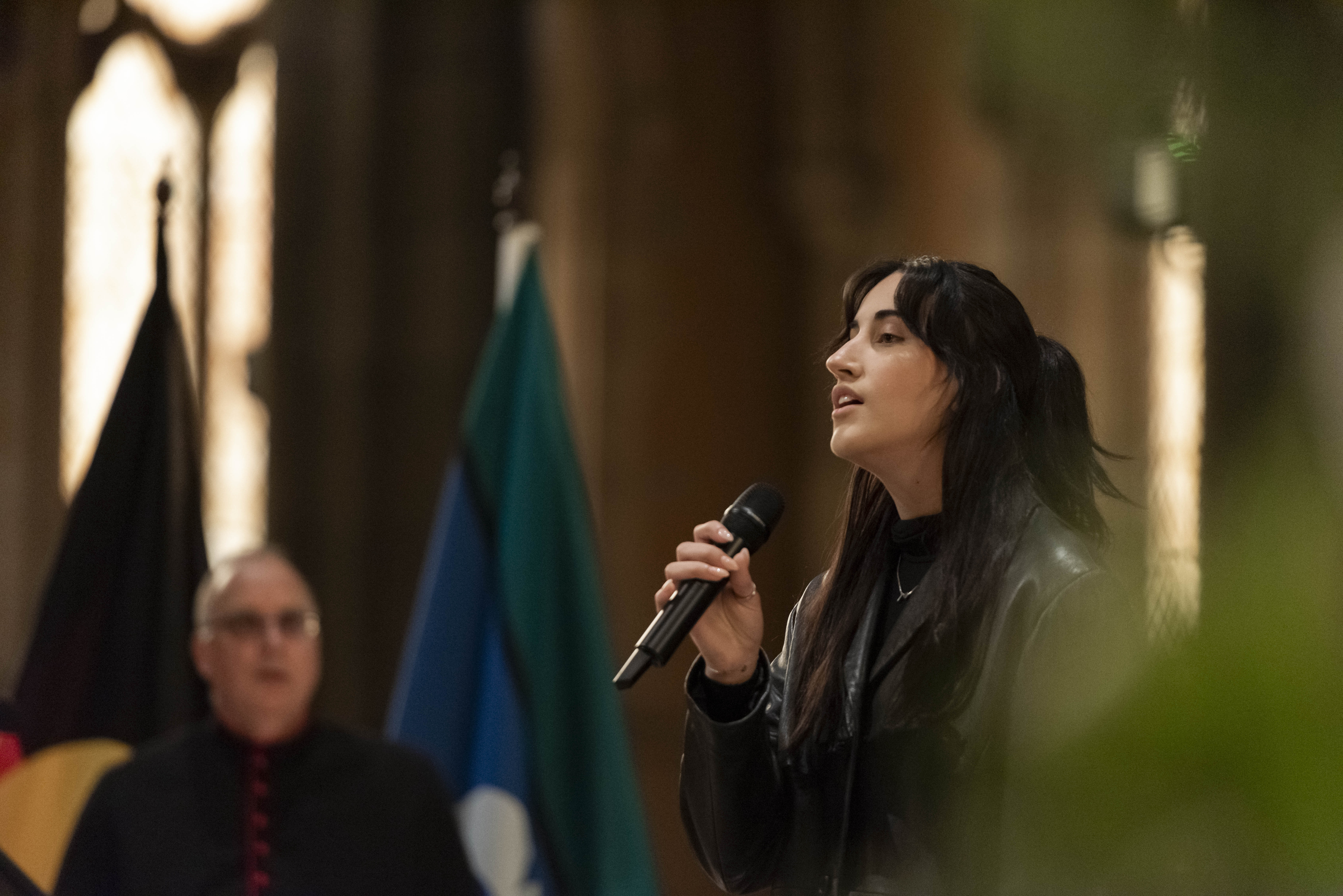 A woman with long black hair in all black clothing sings into a microphone.  